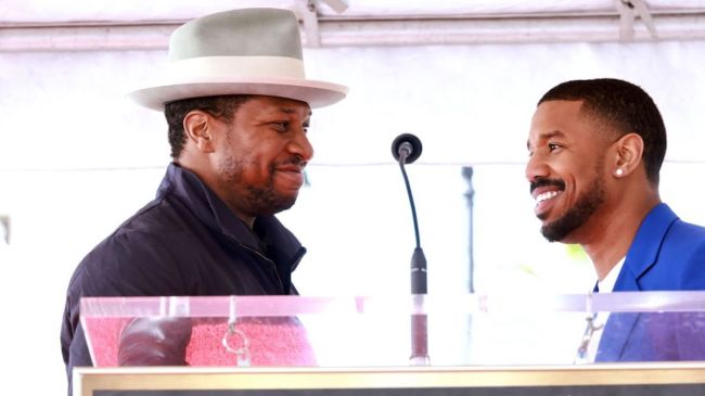 jonathan majors and michael b jordan smiling at each other.