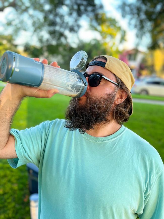 Beard man in sunglasses and a backwards hat holding a Ninja Blast Portable Blender in a city park after making a delicious peach smoothie with it.